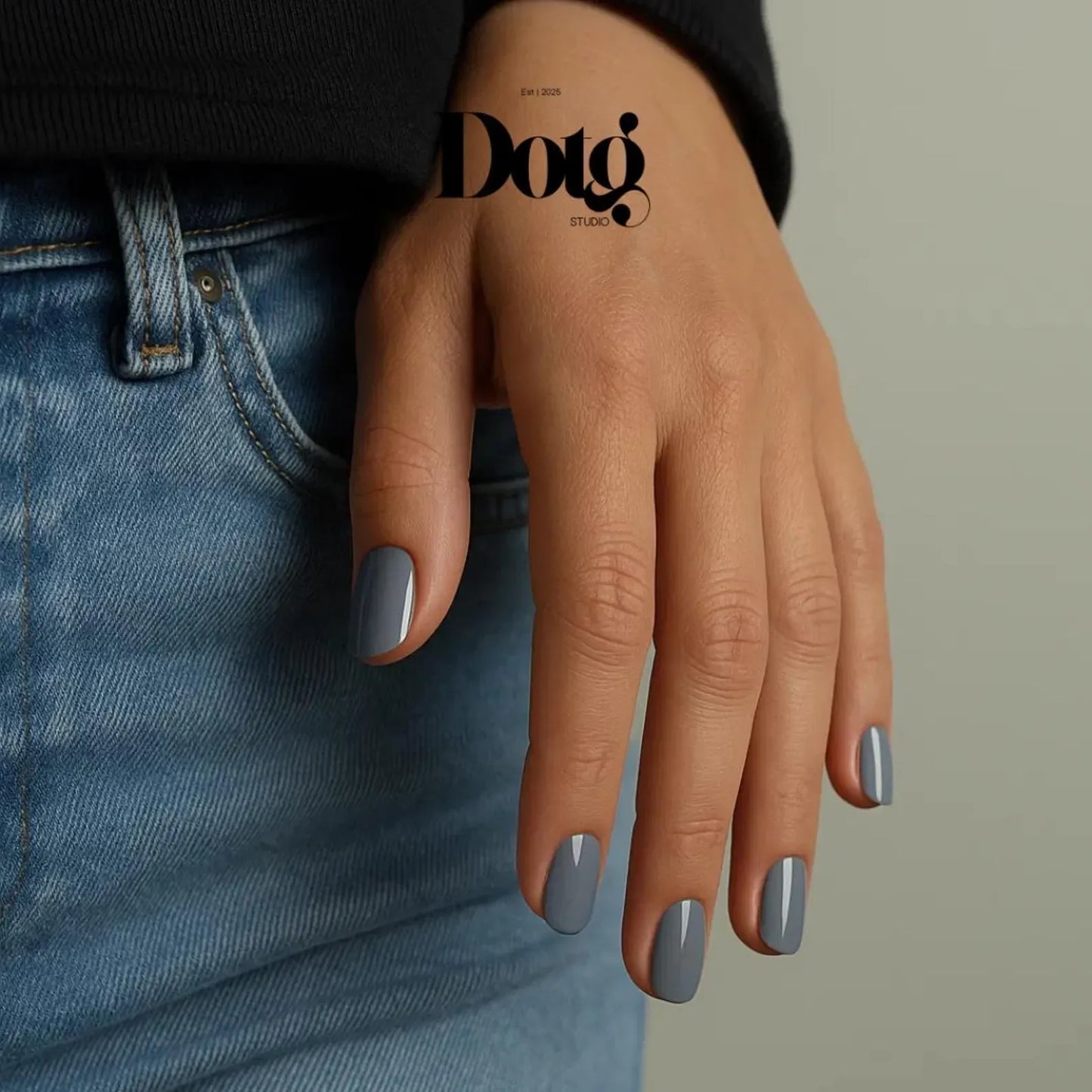 Hand with gray nail polish wearing a black sleeve and blue jeans on a neutral background